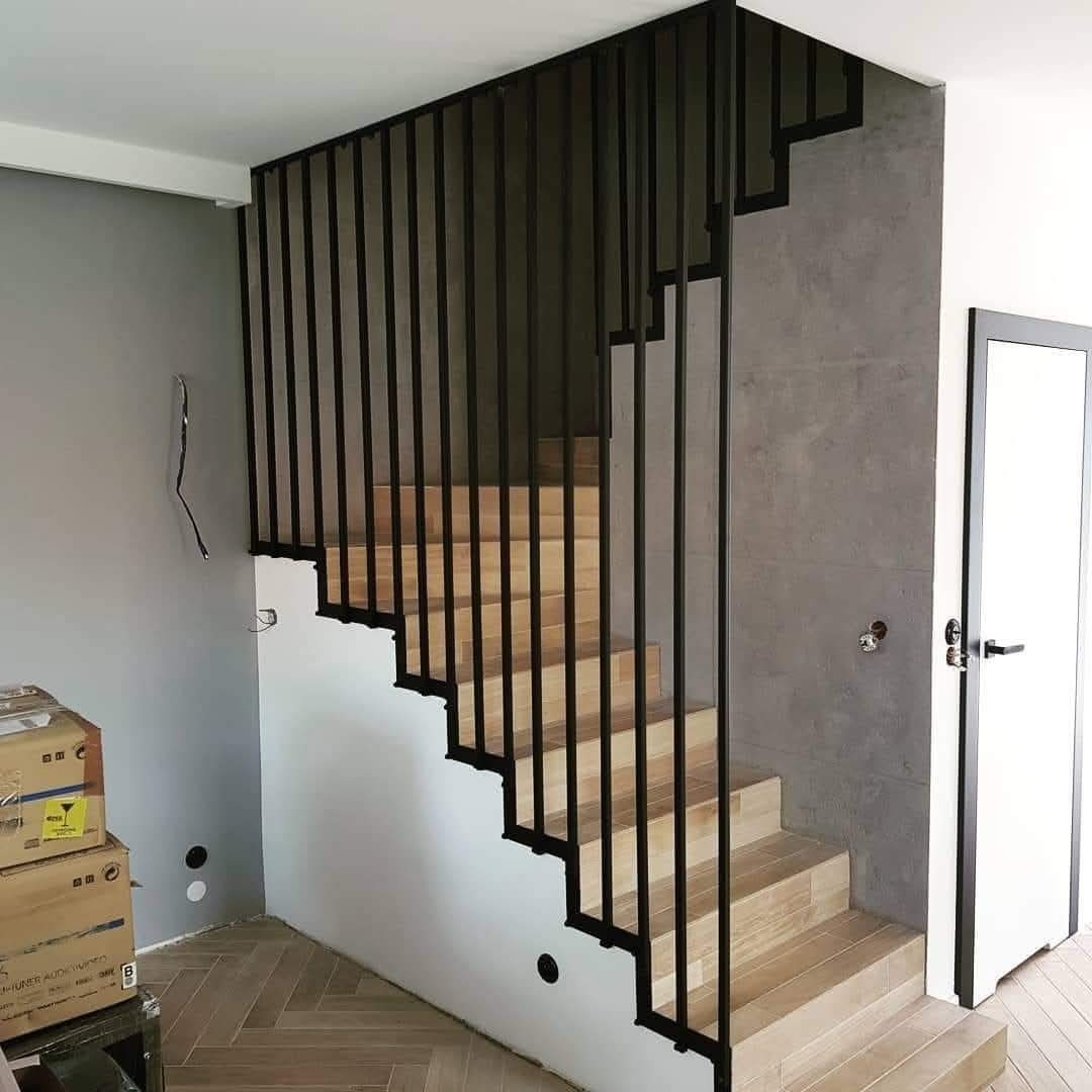Nowoczesne schody z drewnianymi stopniami i czarną, minimalistyczną balustradą z pionowych prętów. Widoczne detale wykończenia wnętrza i podłoga z parkietu.