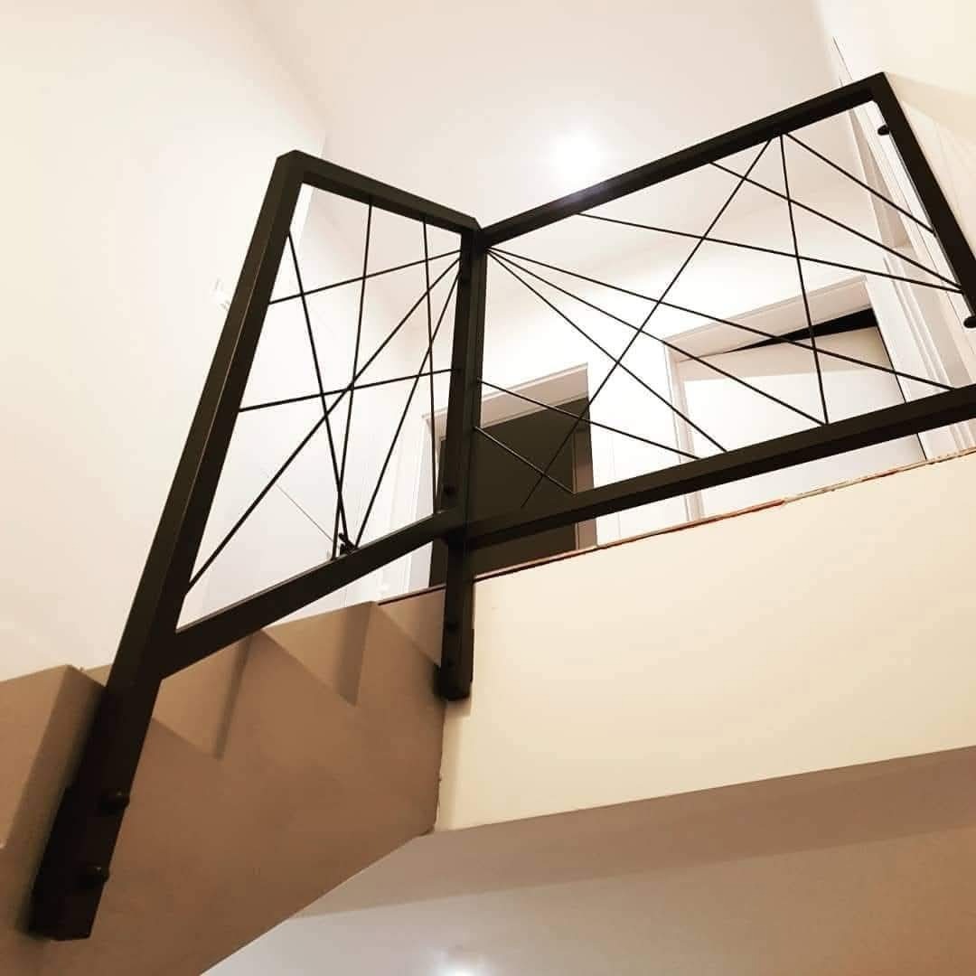 Nowoczesna balustrada schodowa z czarnego metalu o geometrycznym wzorze, widoczna z dołu, z białymi ścianami i drzwiami w tle. Minimalistyczny design.