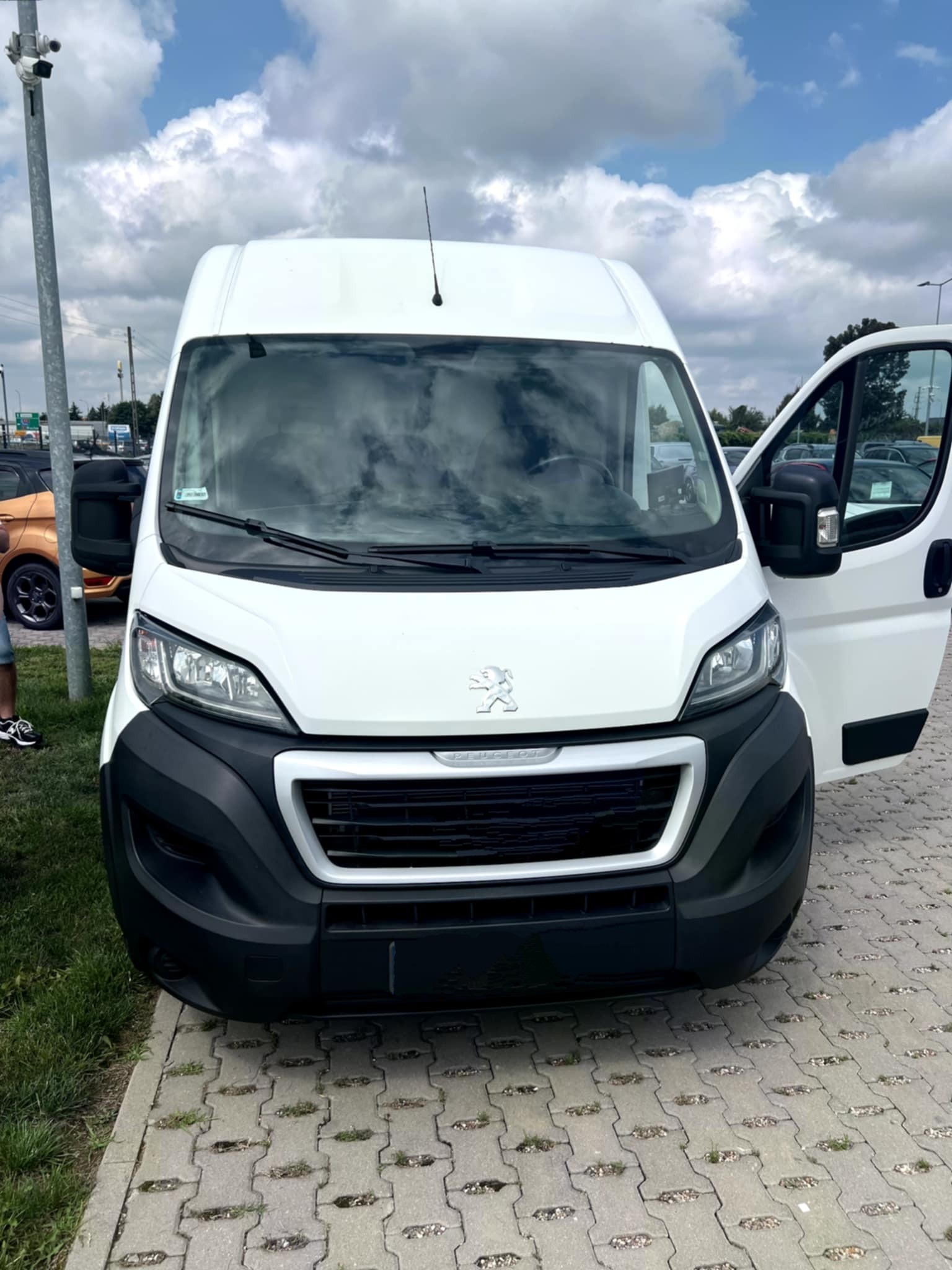 Biały Peugeot Boxer z otwartymi drzwiami na parkingu, widok z przodu, błękitne niebo z chmurami odbija się w szybie.