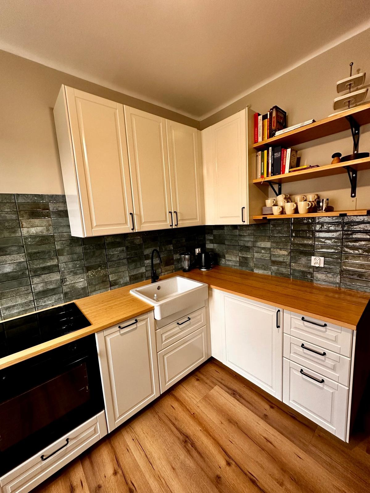 Jasna, narożna kuchnia z drewnianym blatem i ciemnozielonymi płytkami. Uwagę przyciąga ceramiczny zlew i otwarte półki z książkami i filiżankami.