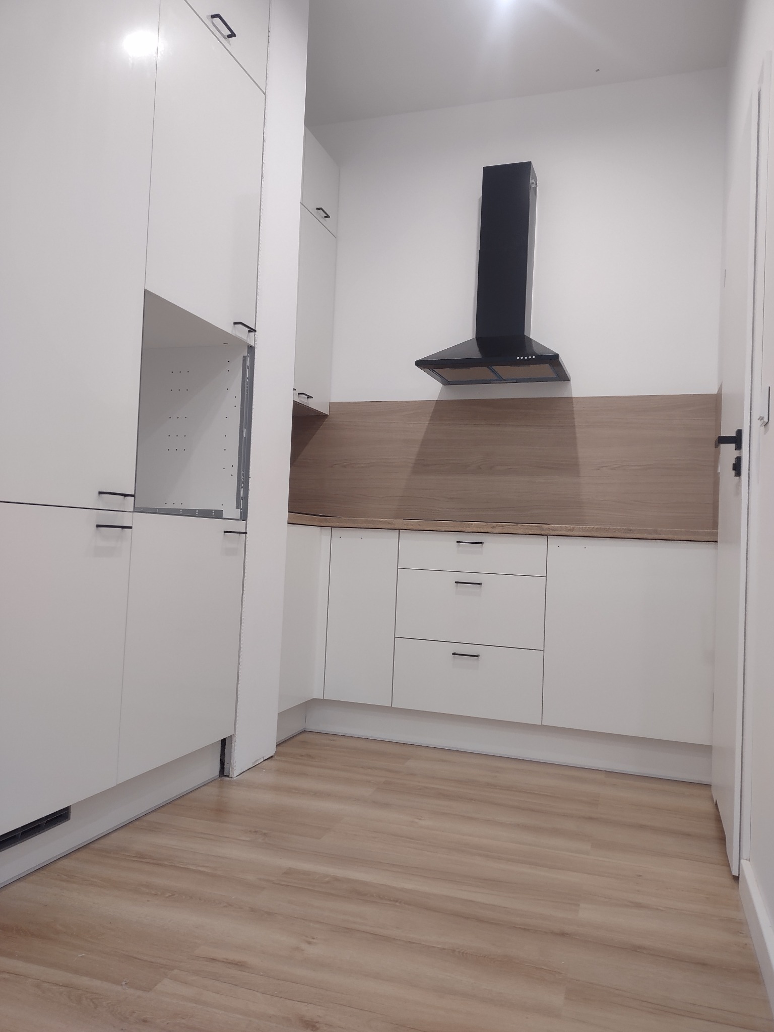 Biała kuchnia z czarnym okapem i panelami drewnopodobnymi. Widoczne szafki, szuflady z minimalistycznymi uchwytami i fragment podłogi z paneli.