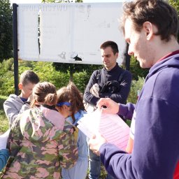 Klaudio - Klaudiusz Włodarczak - Grupa dzieci i dorosłych z kartkami w rękach, prawdopodobnie podczas gry terenowej na świeżym powietrzu, z tablicą informacyjną w tle.