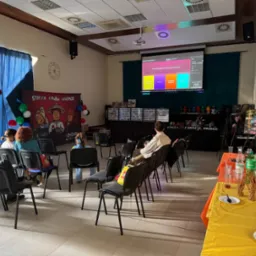 Sala konferencyjna z projekcją quizu tematycznego, dekoracjami i przekąskami, sugerująca imprezę dla dzieci z motywem Ninjago. Uczestnicy siedzą na krzesłach.