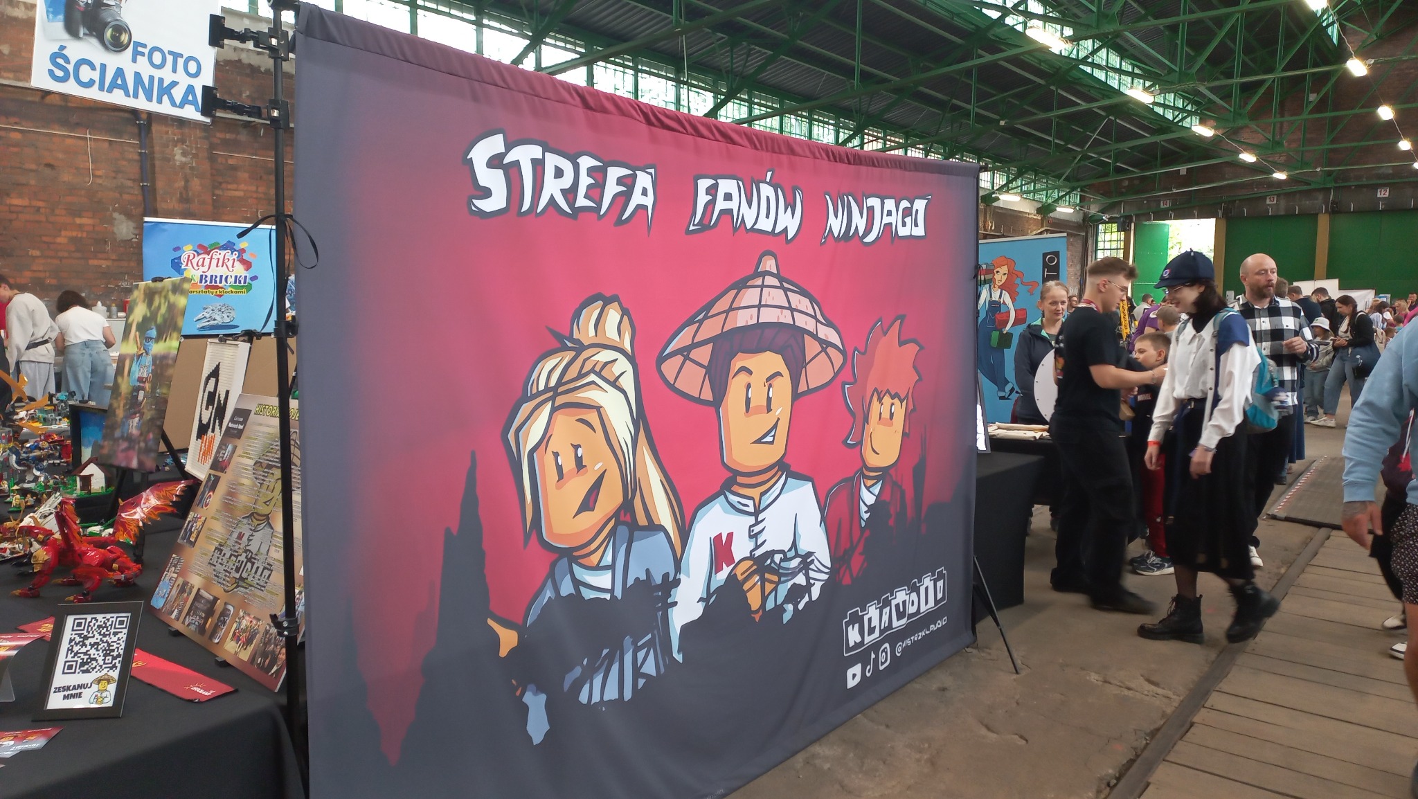 Strefa Fanów Ninjago: ścianka z grafiką postaci z klocków LEGO na tle czerwieni, otoczona tłumem ludzi na evencie. Widoczny napis 'Foto ścianka'.