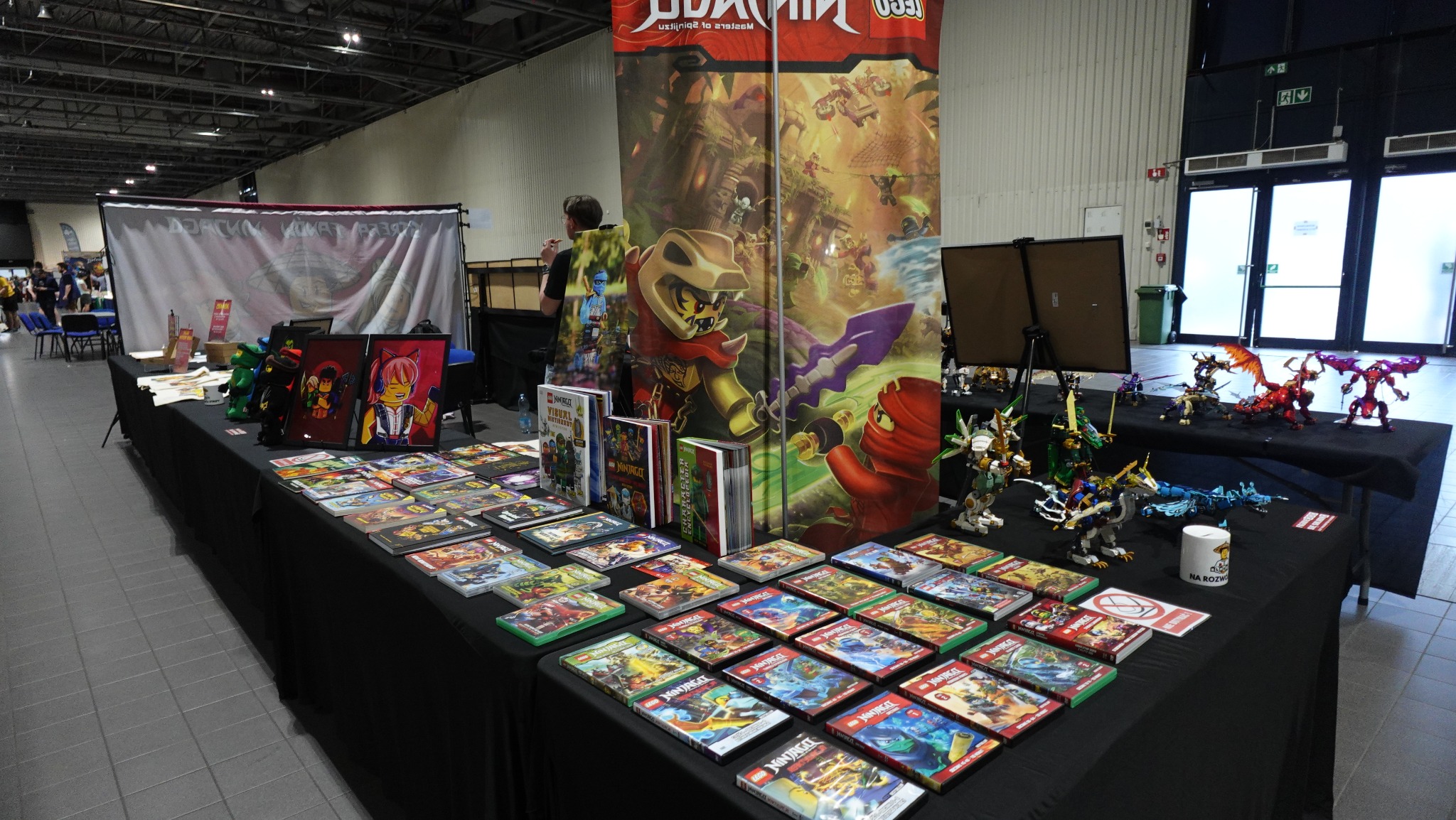 Stoisko z zabawkami Lego Ninjago na evencie w Poznaniu. Widoczne figurki, książki i plakaty z motywem Ninjago. W tle widoczny baner reklamowy.
