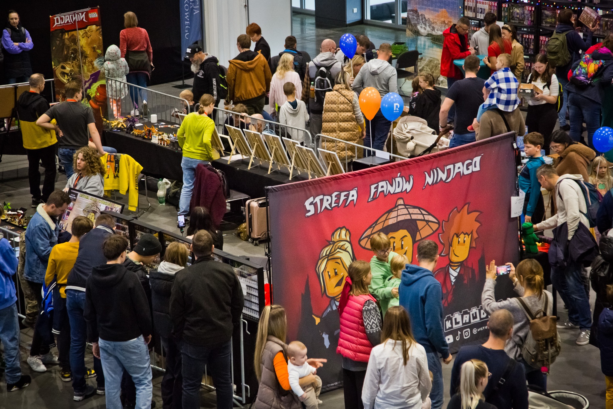 Tłum ludzi na evencie tematycznym LEGO Ninjago, z banerem 'Strefa Fanów Ninjago'. W tle stoły z ekspozycją LEGO i balony. Uczestnicy oglądają wystawę i robią zdjęcia.