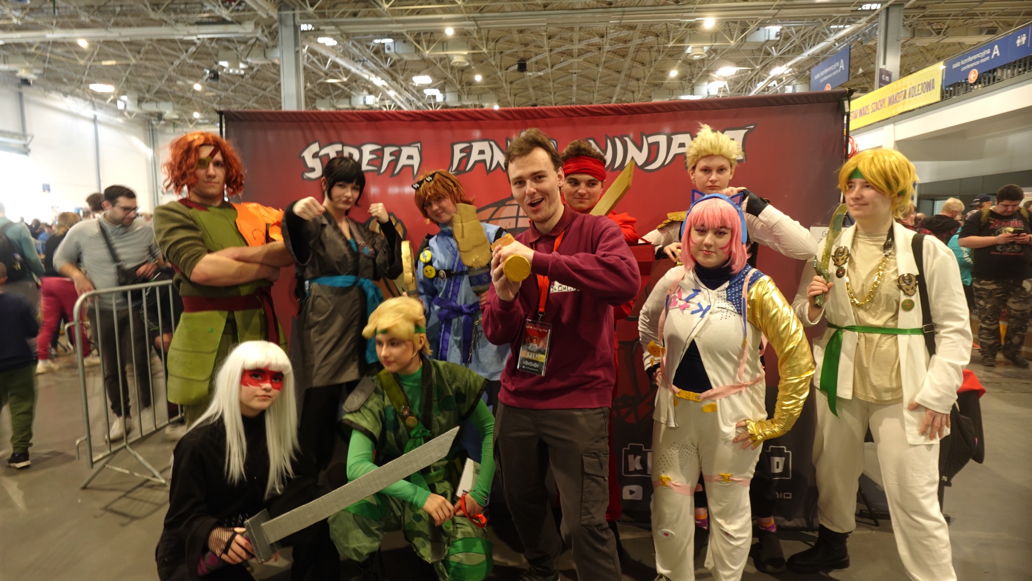 Grupa osób przebranych za postacie z uniwersum NinjaGo pozuje na tle baneru 'Strefa Fan Ninja'. Uczestnicy w kostiumach, z rekwizytami, na konwencie.