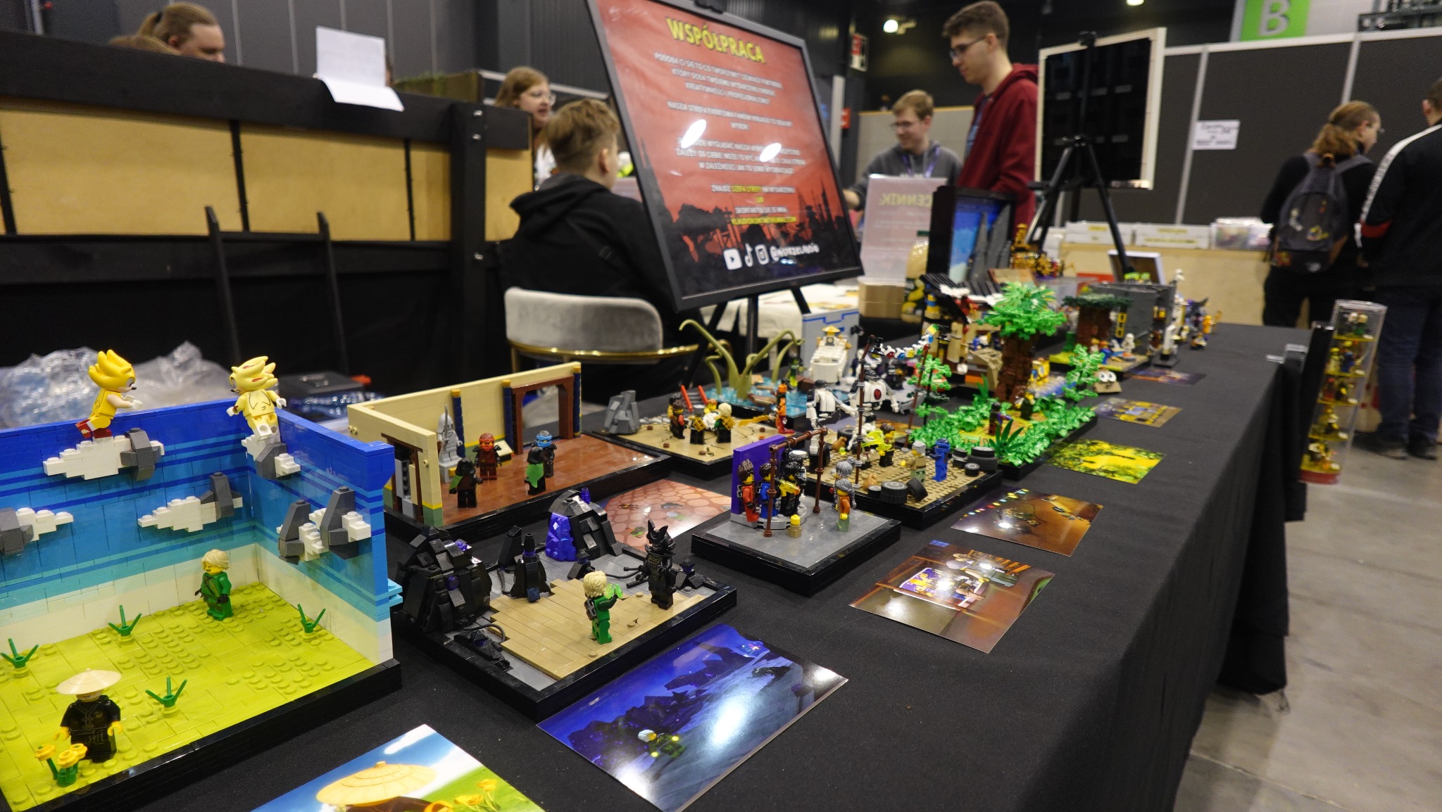 Stoisko z ekspozycją dioram z klocków Lego, prezentujących sceny walki i krajobrazy, na tle osób i plakatu 'Współpraca' podczas wydarzenia.