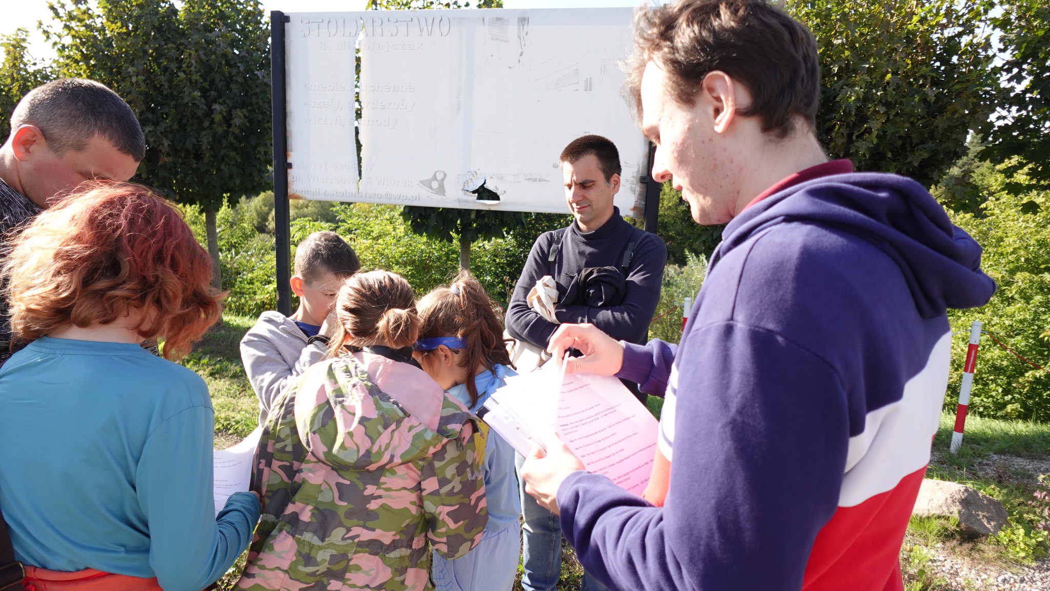 Grupa dzieci i dorosłych z kartkami w rękach, prawdopodobnie podczas gry terenowej na świeżym powietrzu, z tablicą informacyjną w tle.