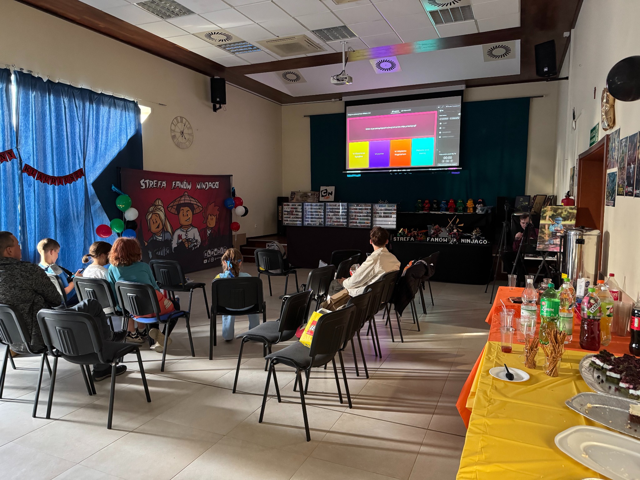Sala konferencyjna z projekcją quizu tematycznego, dekoracjami i przekąskami, sugerująca imprezę dla dzieci z motywem Ninjago. Uczestnicy siedzą na krzesłach.