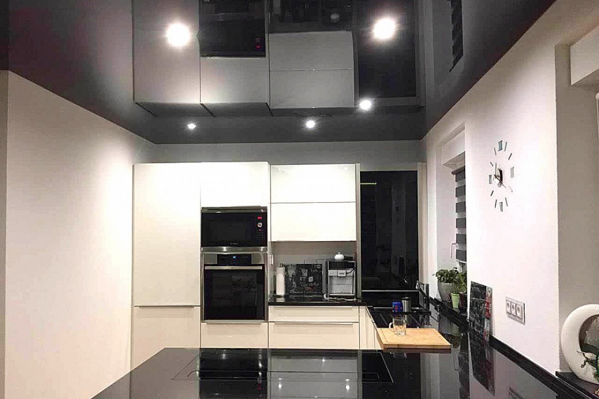 Nowoczesna kuchnia z czarnym, błyszczącym sufitem, który odbija światło i meble. Widoczne białe szafki, sprzęt AGD i minimalistyczny zegar na ścianie.