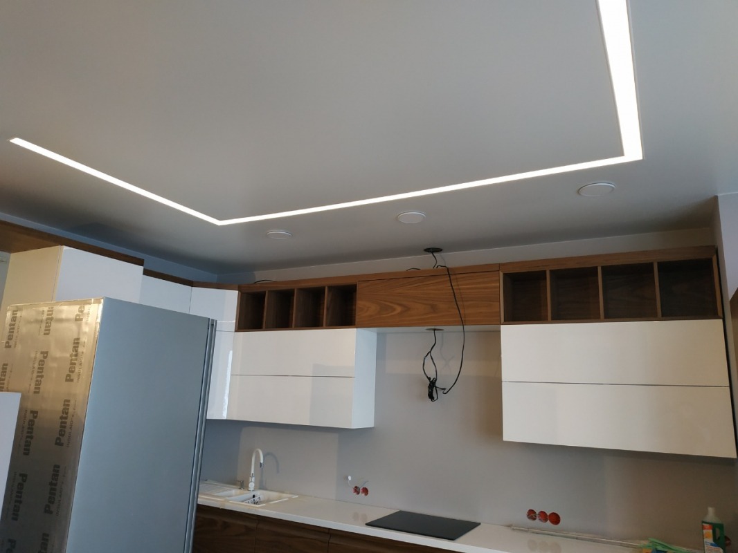 Nowoczesny sufit napinany z oświetleniem LED w kształcie litery L, wkomponowany w aranżację kuchni z białymi szafkami i drewnianymi akcentami.