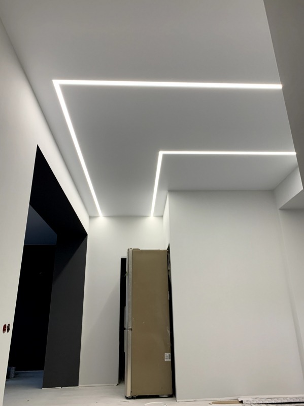 Białe wnętrze z nowoczesnym oświetleniem LED w kształcie litery L na suficie. Widoczny fragment korytarza i lodówka. Minimalistyczny design, jasne kolory.