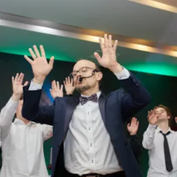 Wokalista z mikrofonem nagłownym i grupa osób z podniesionymi rękami podczas występu w Ostrowie Wielkopolskim, oświetlenie sceniczne.