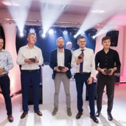 Wręczenie nagród na imprezie firmowej: pięciu mężczyzn w eleganckich strojach trzyma drewniane pudełka na tle oświetlonej sceny, konferansjer w smokingu z mikrofonem.