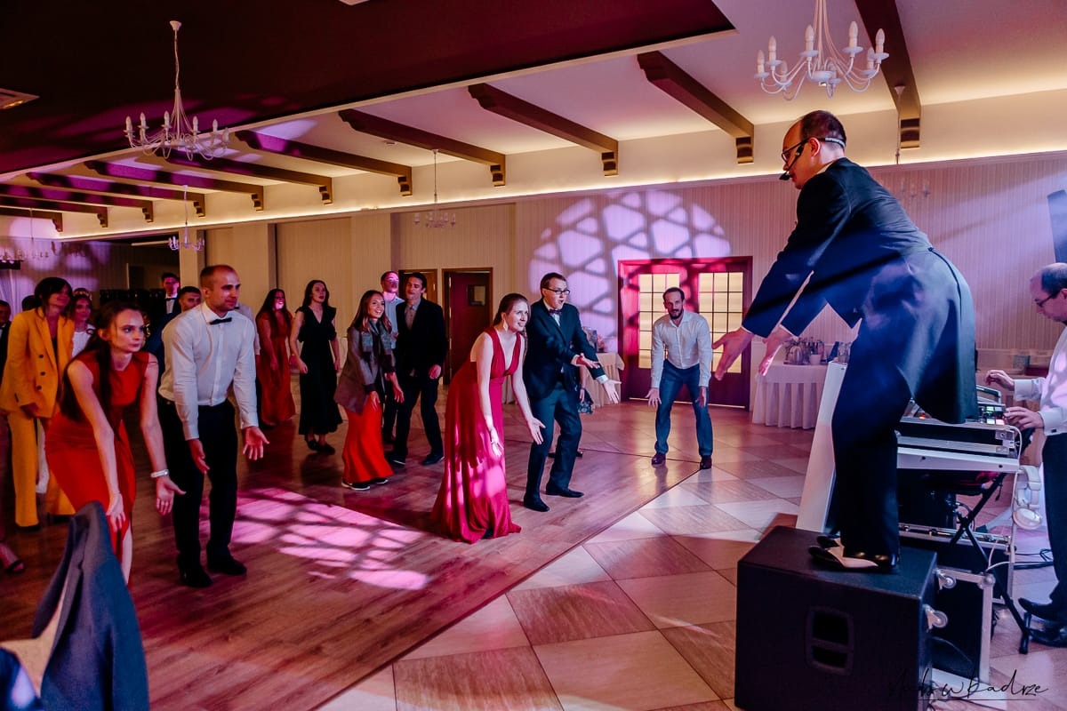 Dj na podwyższeniu prowadzi animacje taneczne z gośćmi weselnymi w sali balowej z parkietem w kratkę i oświetleniem w odcieniach fioletu i różu.