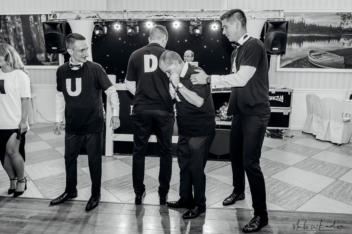 Czarno-białe zdjęcie z Ostrów Wielkopolski przedstawia czterech mężczyzn w czarnych koszulkach z białymi literami U i D, stojących na scenie z oświetleniem i dekoracjami, gdzie jeden z nich jest...