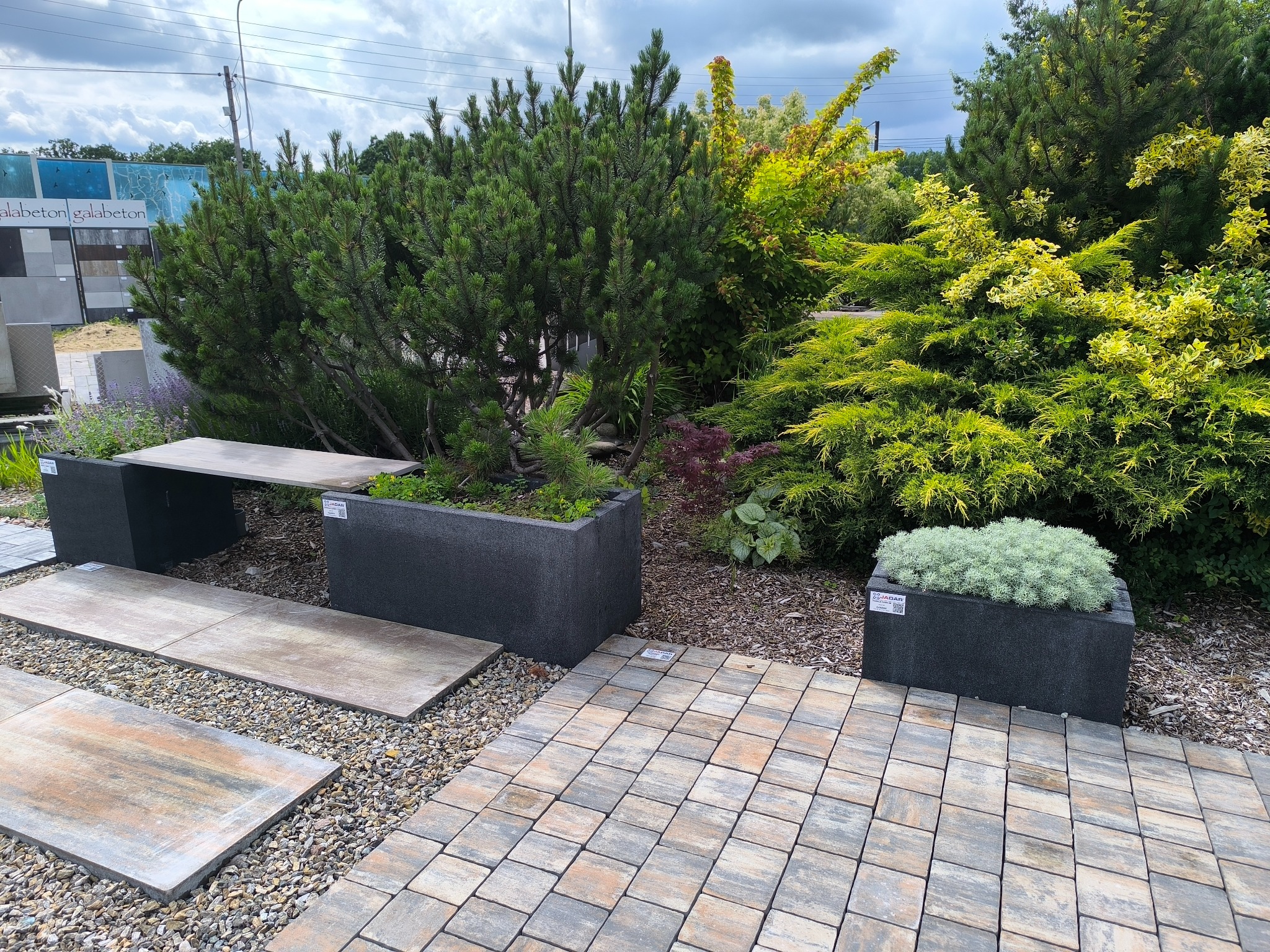 Aranżacja ogrodu z kostki brukowej, donic i roślin. Ekspozycja materiałów budowlanych Galabeton. Widoczne różne tekstury i kolory, tworzące harmonijną przestrzeń.