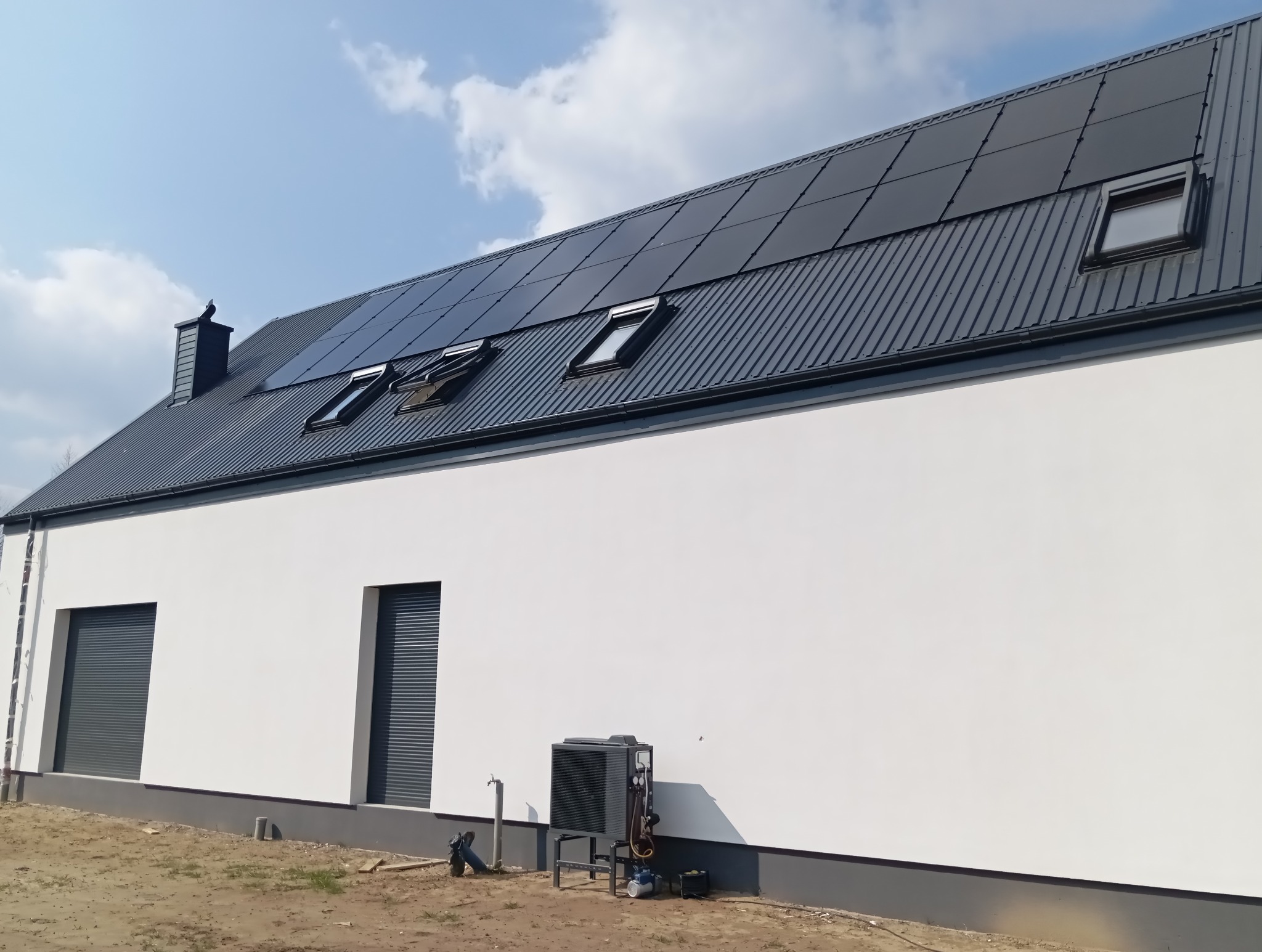 Dom z panelami fotowoltaicznymi na dachu, oknami dachowymi i pompą ciepła przy białej ścianie. Widok na kompletną instalację z zewnątrz.