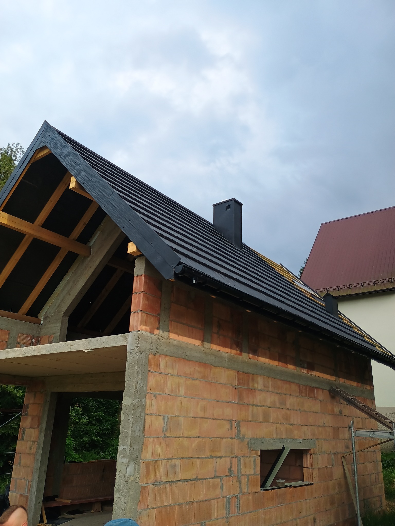 Ceglany budynek w trakcie rozbudowy z czarnym dachem i kominem, widoczne drewniane elementy konstrukcyjne. W tle pochmurne niebo.