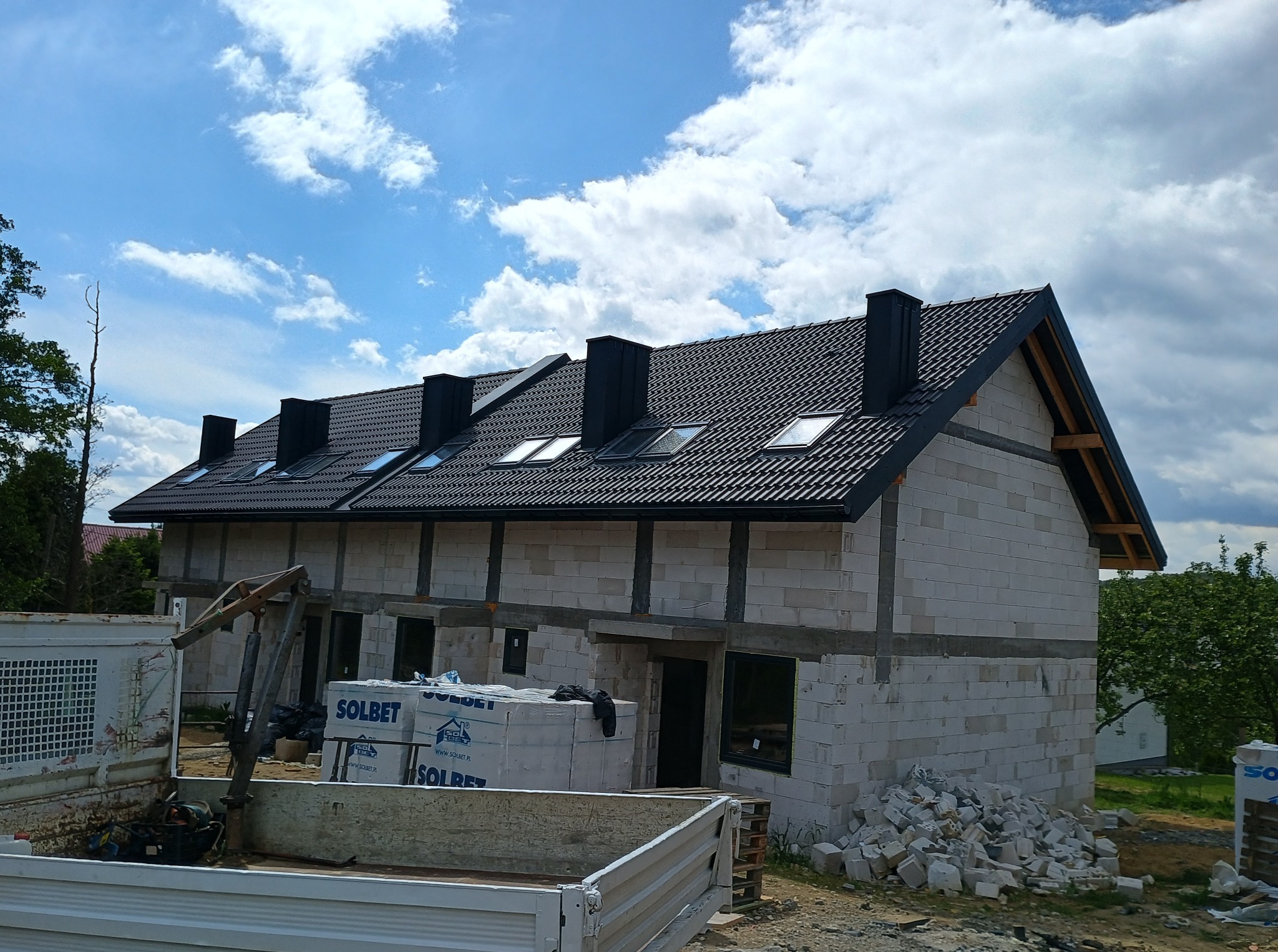 Rozbudowa bliźniaka w stanie surowym otwartym z czarną dachówką, okna dachowe, białe bloczki, palety z materiałami budowlanymi i fragment wywrotki na pierwszym planie.