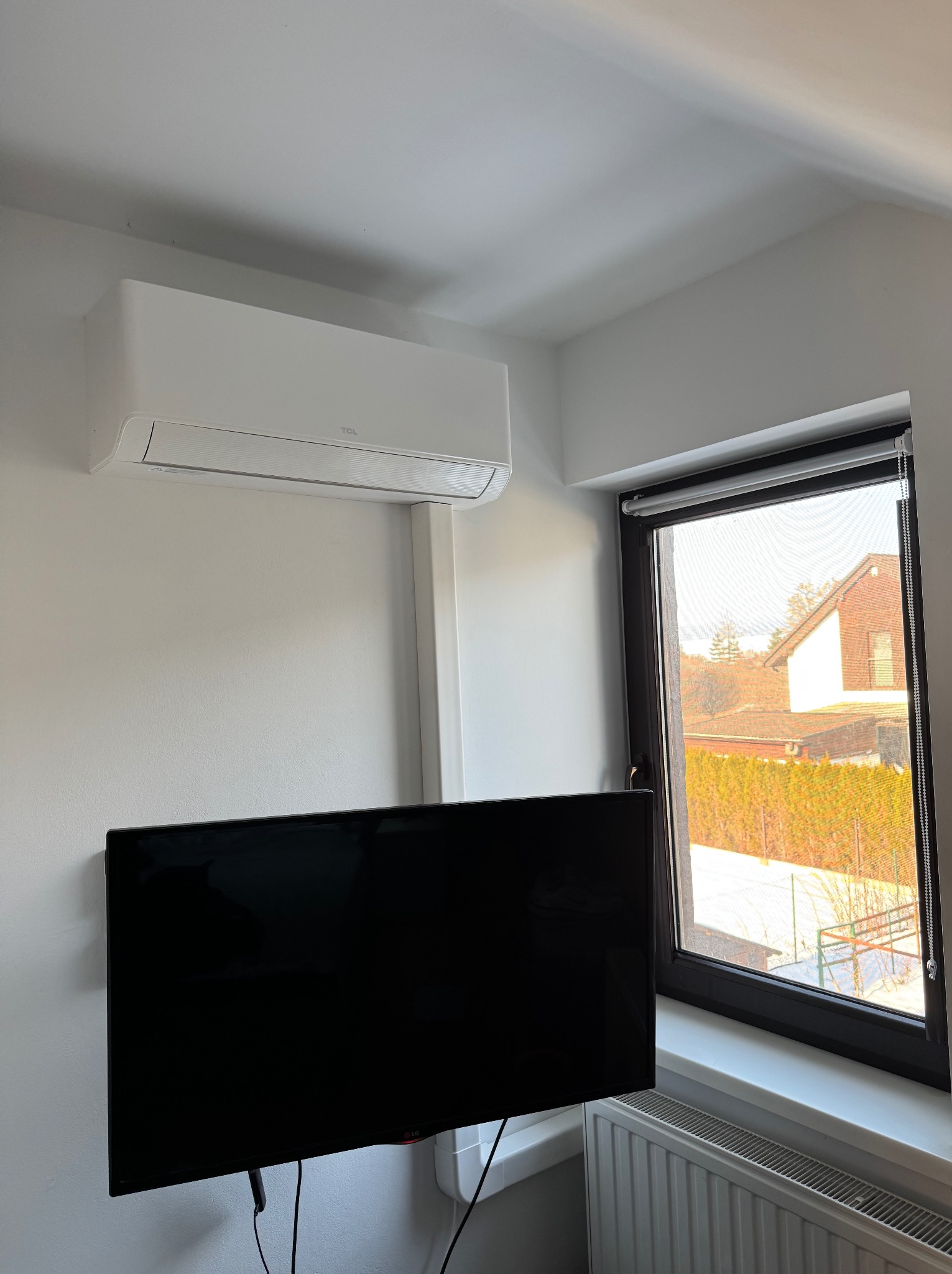 Biały klimatyzator TCL zamontowany na ścianie w pokoju, obok okna z widokiem na dom i ogród. Pod klimatyzatorem wisi czarny telewizor.