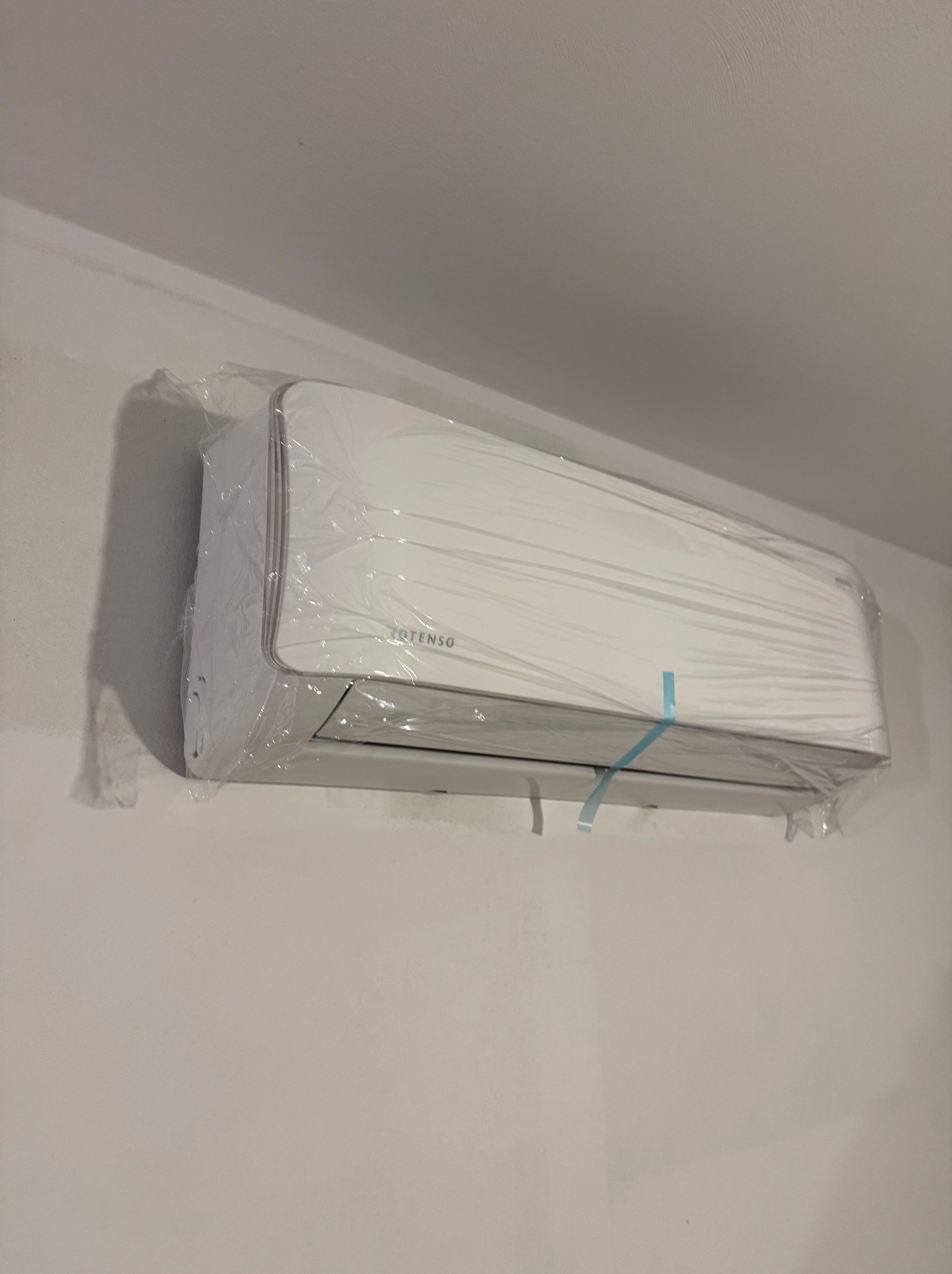 Biała jednostka klimatyzacji marki Totenso, jeszcze w folii ochronnej, zamontowana na jasnej ścianie. Widoczne delikatne niedoskonałości ściany.