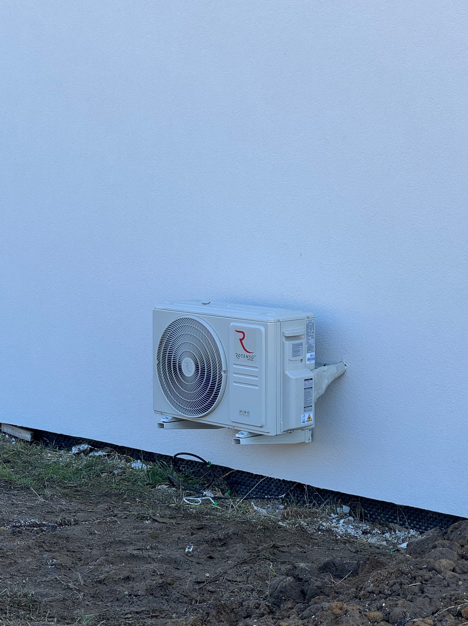 Biała jednostka zewnętrzna klimatyzacji Rotenso zamontowana na jasnej ścianie budynku. Widoczne przewody i fragment trawnika u dołu kadru.