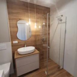 Nowoczesna łazienka z drewnopodobnymi płytkami, asymetrycznym lustrem i kabiną prysznicową ze szkła. Minimalistyczny design, ciepłe oświetlenie, jasne barwy.