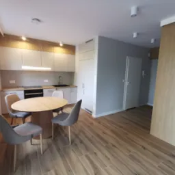 Jasna kuchnia z drewnianymi akcentami: okrągły stół z szarymi krzesłami, zabudowa kuchenna z oświetleniem LED, podłoga imitująca drewno, minimalistyczny wystrój.