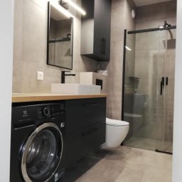 PRODESIGN SPÓŁKA Z OGRANICZONĄ ODPOWIEDZIALNOŚCIĄ - Nowoczesna łazienka z czarnymi akcentami: pralka, szafka pod umywalką, wysoka szafka wisząca. Kabina prysznicowa ze szklanymi drzwiami. Jasne płytki na ścianach i podłodze.