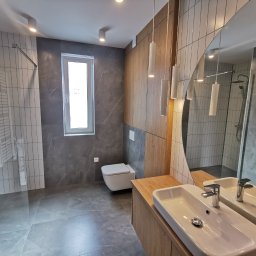 PRODESIGN SPÓŁKA Z OGRANICZONĄ ODPOWIEDZIALNOŚCIĄ - Nowoczesna łazienka po remoncie: szare płytki imitujące marmur, drewniane akcenty, okrągłe lustro i podwieszana toaleta. Widoczny prysznic typu walk-in.