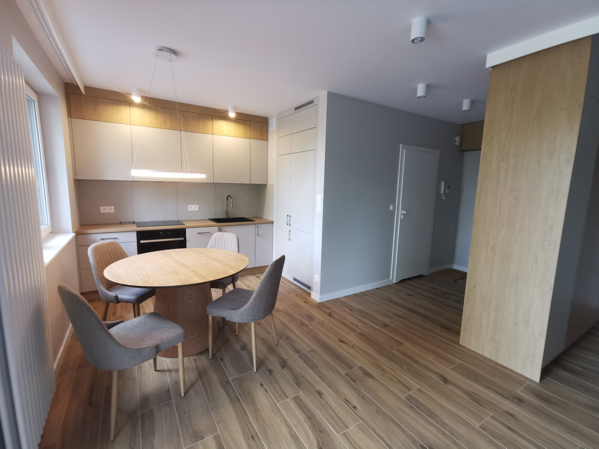 Jasna kuchnia z drewnianymi akcentami: okrągły stół z szarymi krzesłami, zabudowa kuchenna z oświetleniem LED, podłoga imitująca drewno, minimalistyczny wystrój.