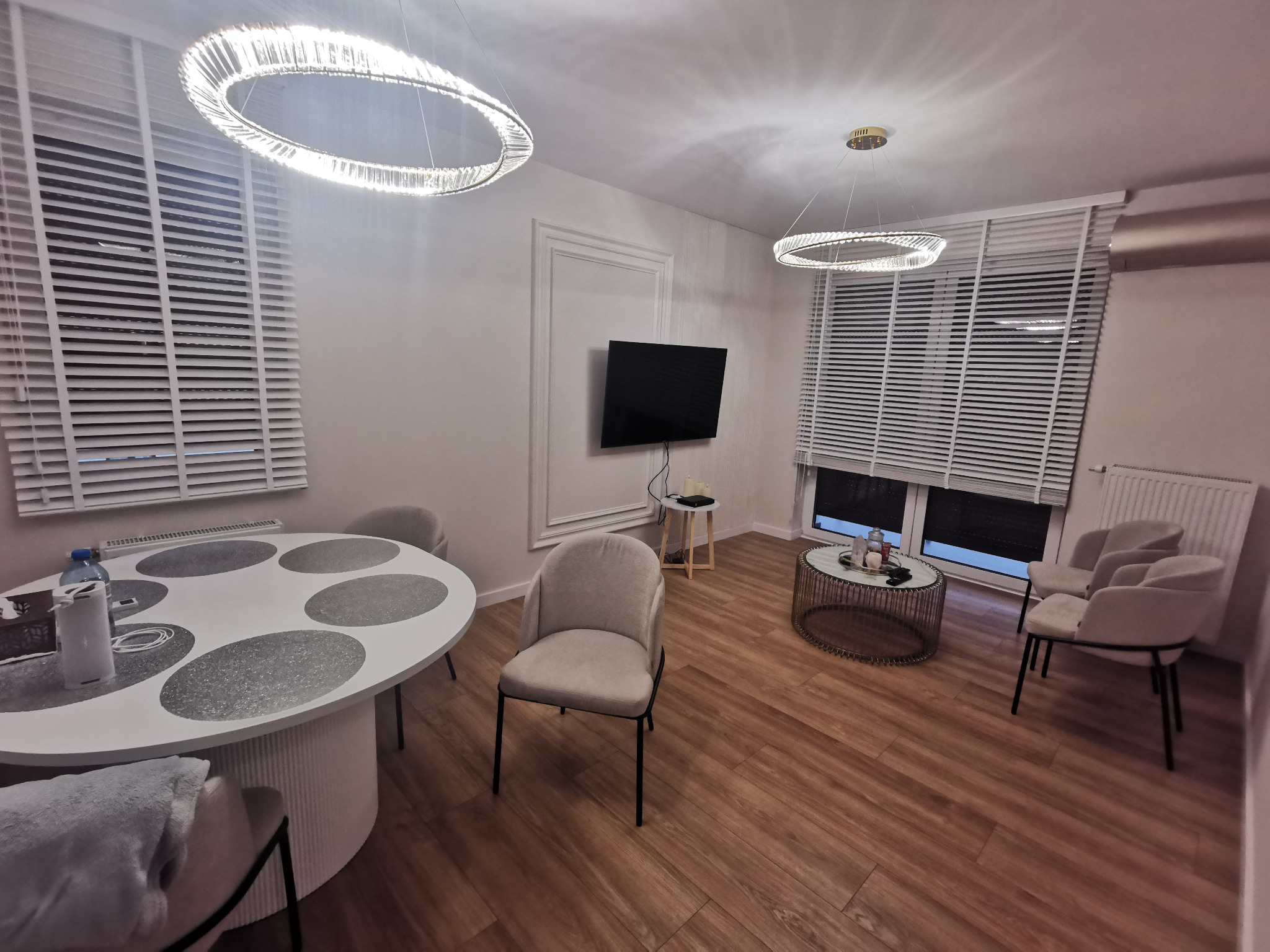 Jasne wnętrze pokoju z drewnianą podłogą, okrągłym stołem i krzesłami. Dwa okrągłe żyrandole. Telewizor na ścianie. Rolety w oknach.