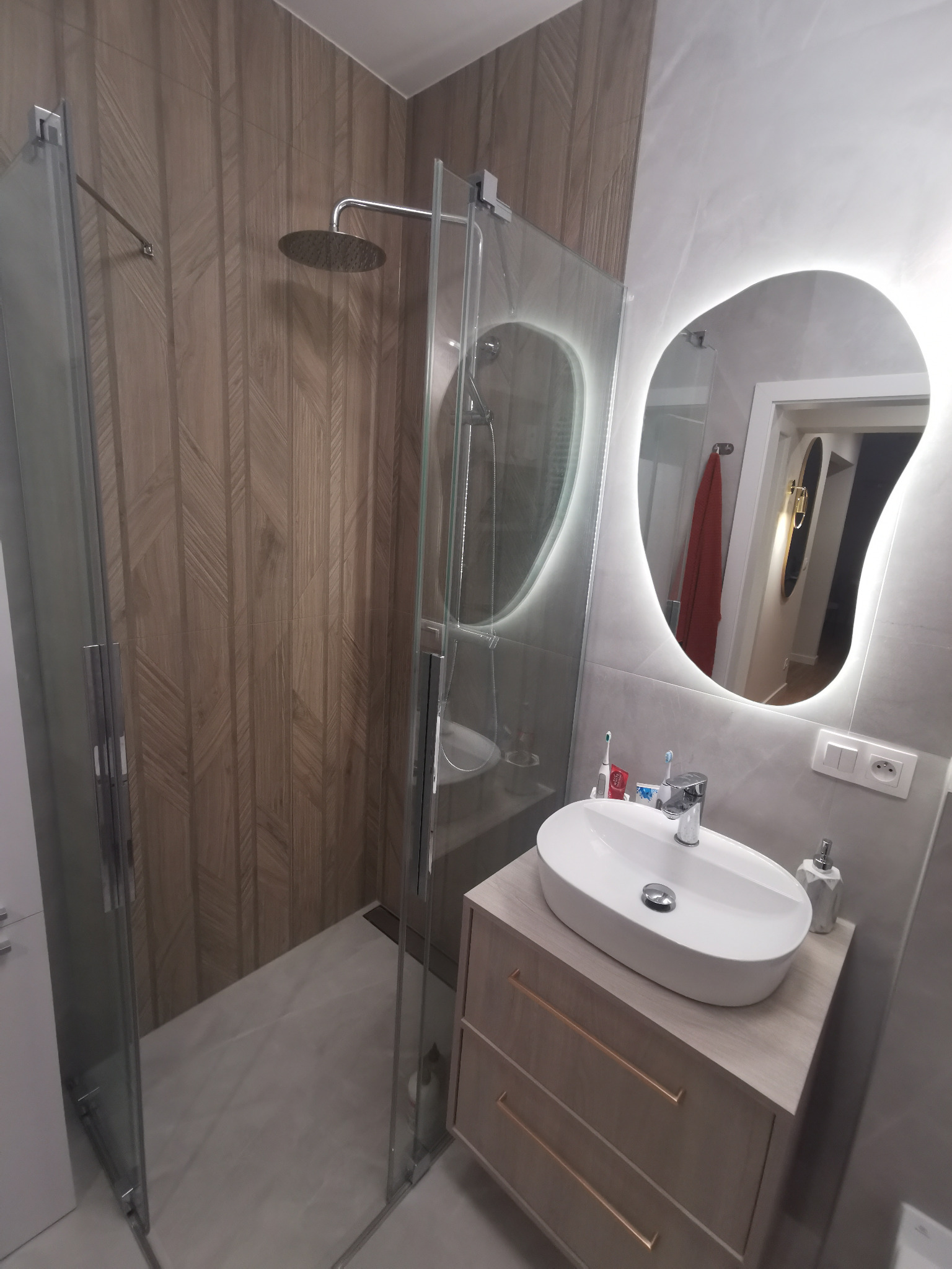 Nowoczesna łazienka po remoncie: kabina prysznicowa ze szklanymi drzwiami, umywalka nablatowa na szafce z szufladami i podświetlane lustro owalne.