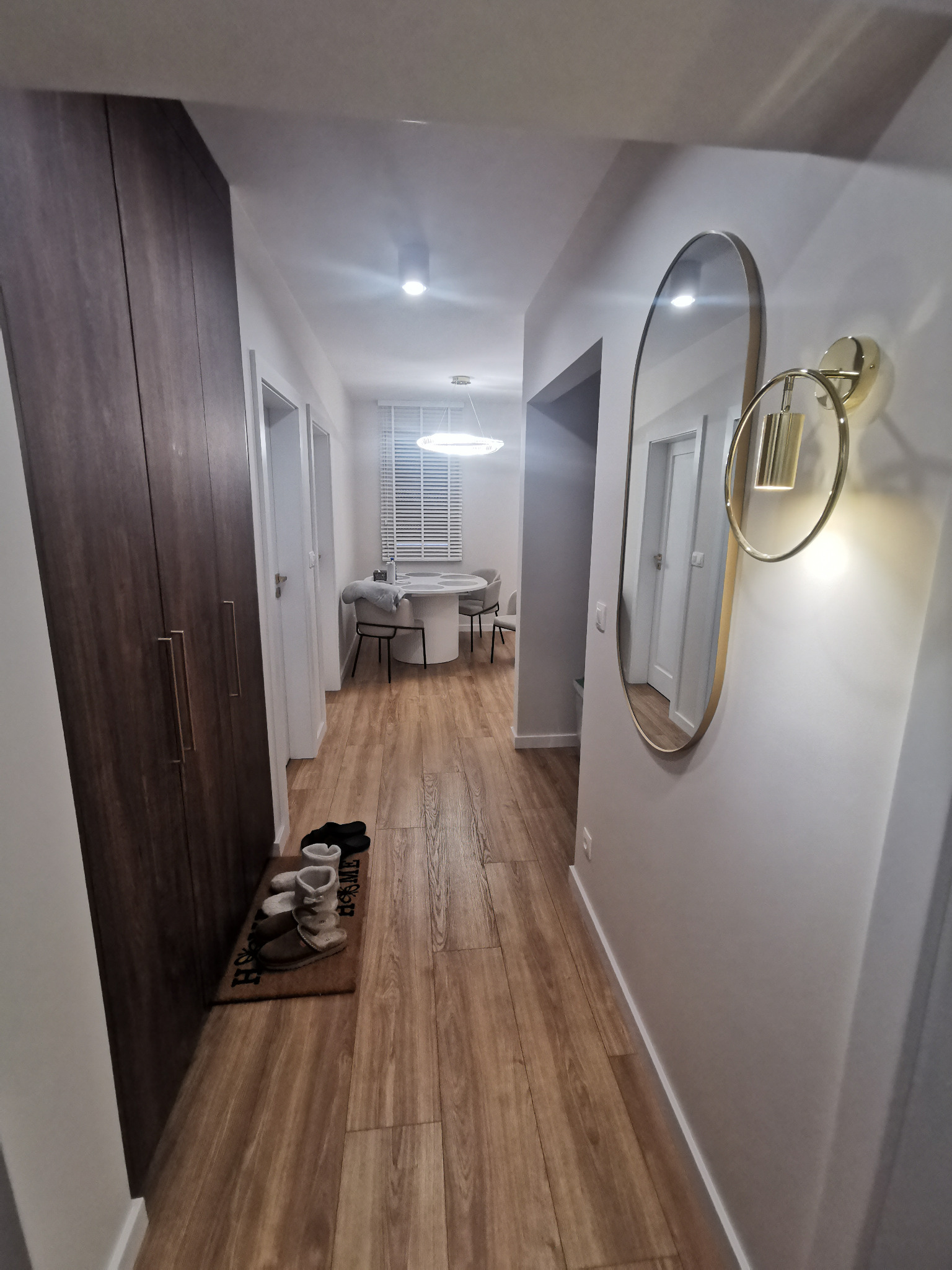 Nowoczesny przedpokój z drewnianą podłogą, szafą w zabudowie i owalnym lustrem z oświetleniem. W tle widoczny stół z krzesłami i rolety w oknie.