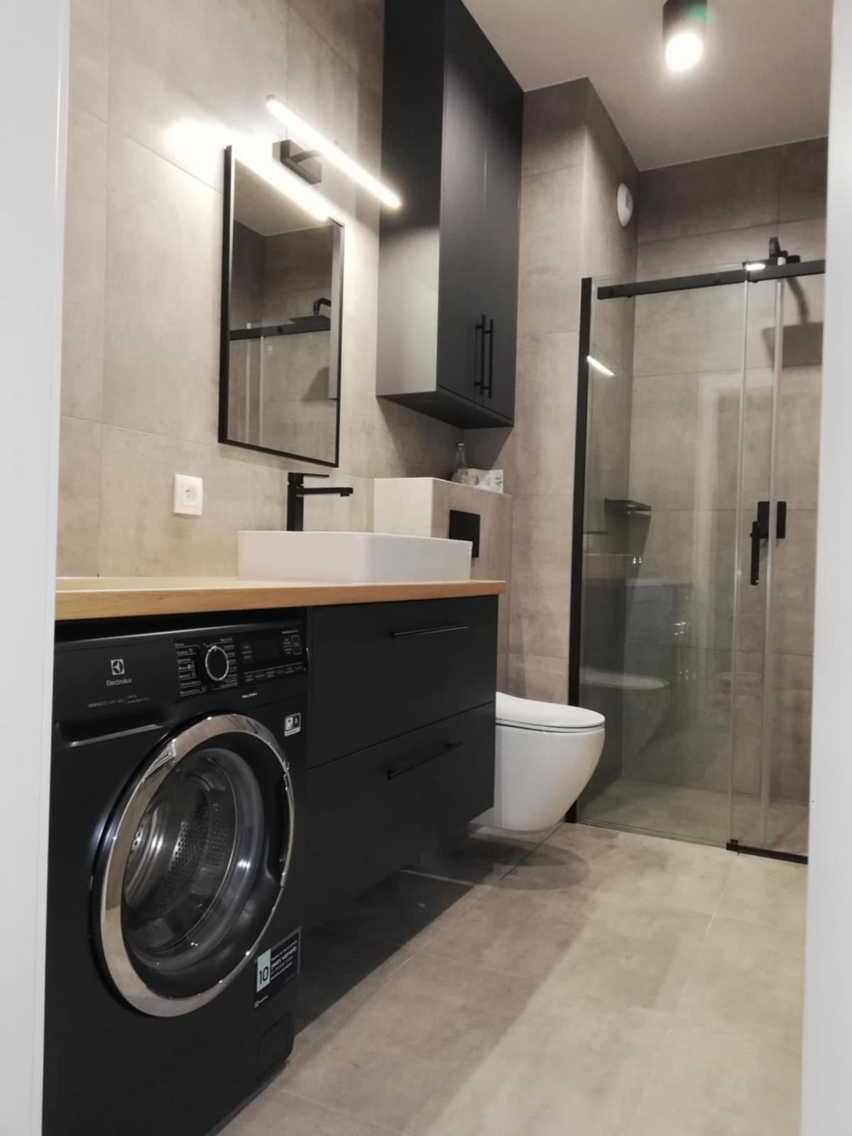 Nowoczesna łazienka z czarnymi akcentami: pralka, szafka pod umywalką, wysoka szafka wisząca. Kabina prysznicowa ze szklanymi drzwiami. Jasne płytki na ścianach i podłodze.