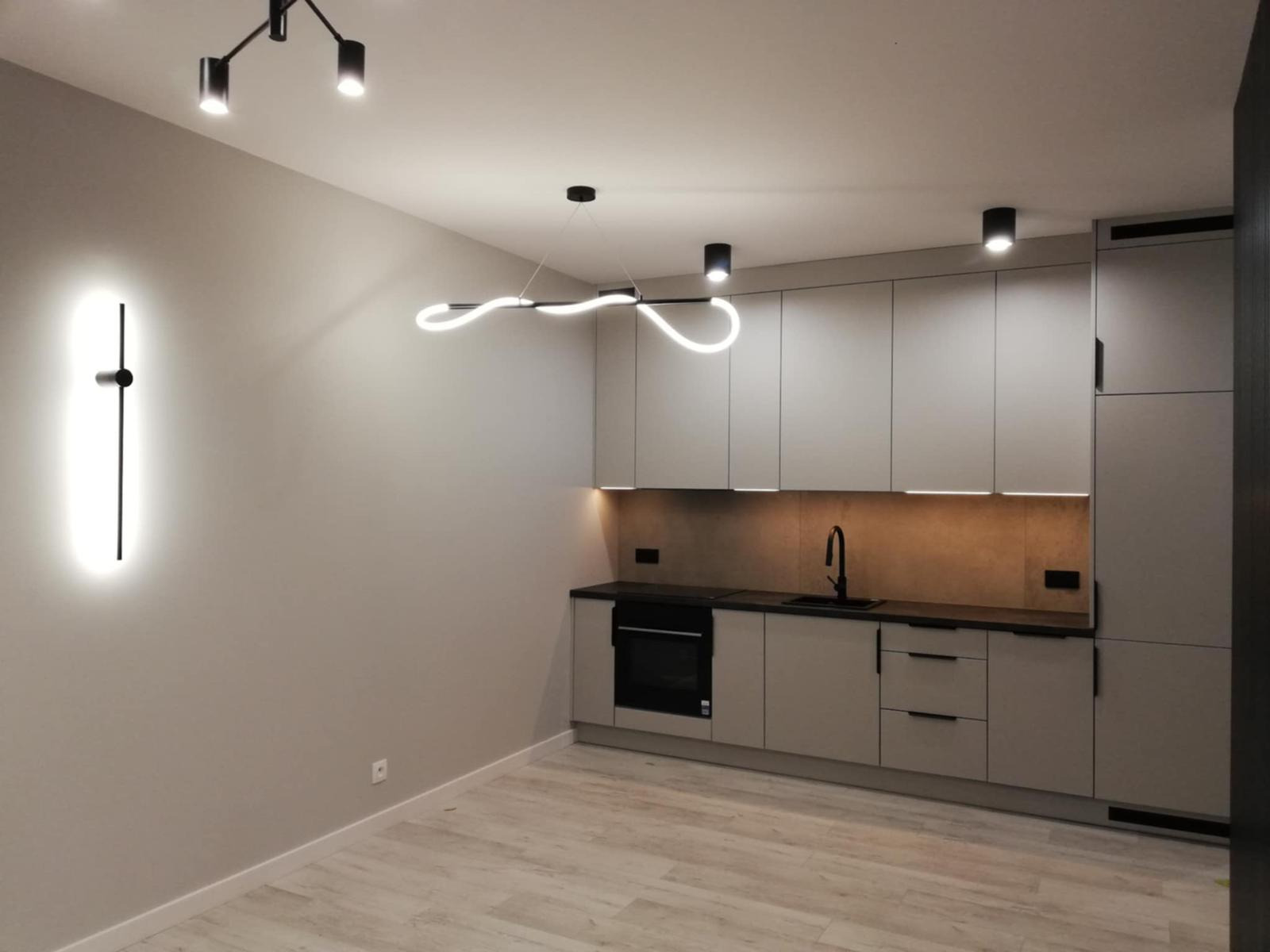 Nowoczesna kuchnia w odcieniach szarości z oryginalnym oświetleniem LED: lampa ścienna i wisząca. Minimalistyczny design, jasne drewniane panele podłogowe.