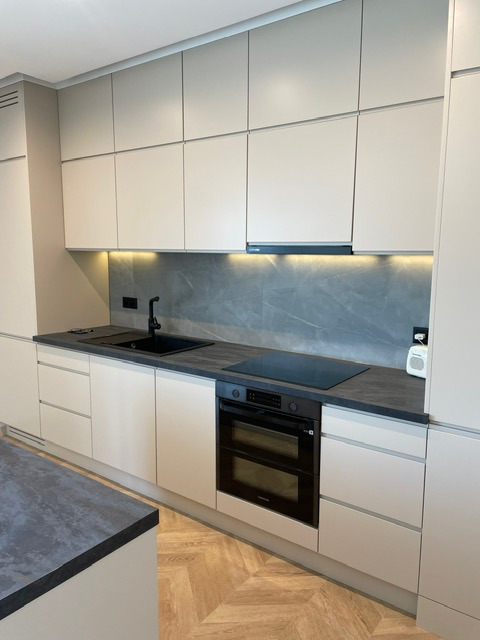 Nowoczesna, jasna kuchnia z szarym blatem i czarnym zlewem. Minimalistyczne szafki, oświetlenie LED pod szafkami. Parkiet ułożony w jodełkę.