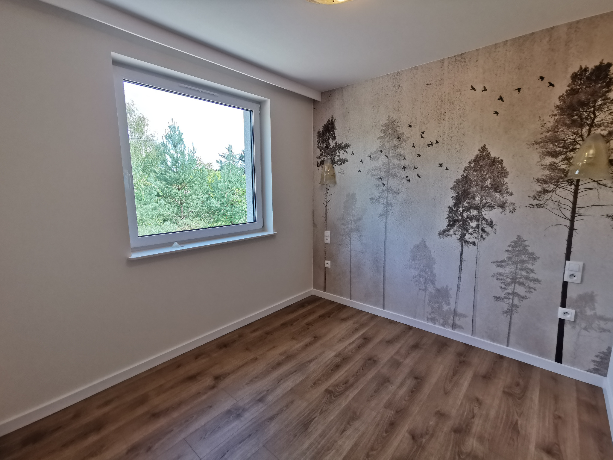 Jasne wnętrze pokoju z drewnianą podłogą i fototapetą z motywem leśnym na ścianie. Widoczne okno z widokiem na zieleń. Minimalistyczny wystrój.