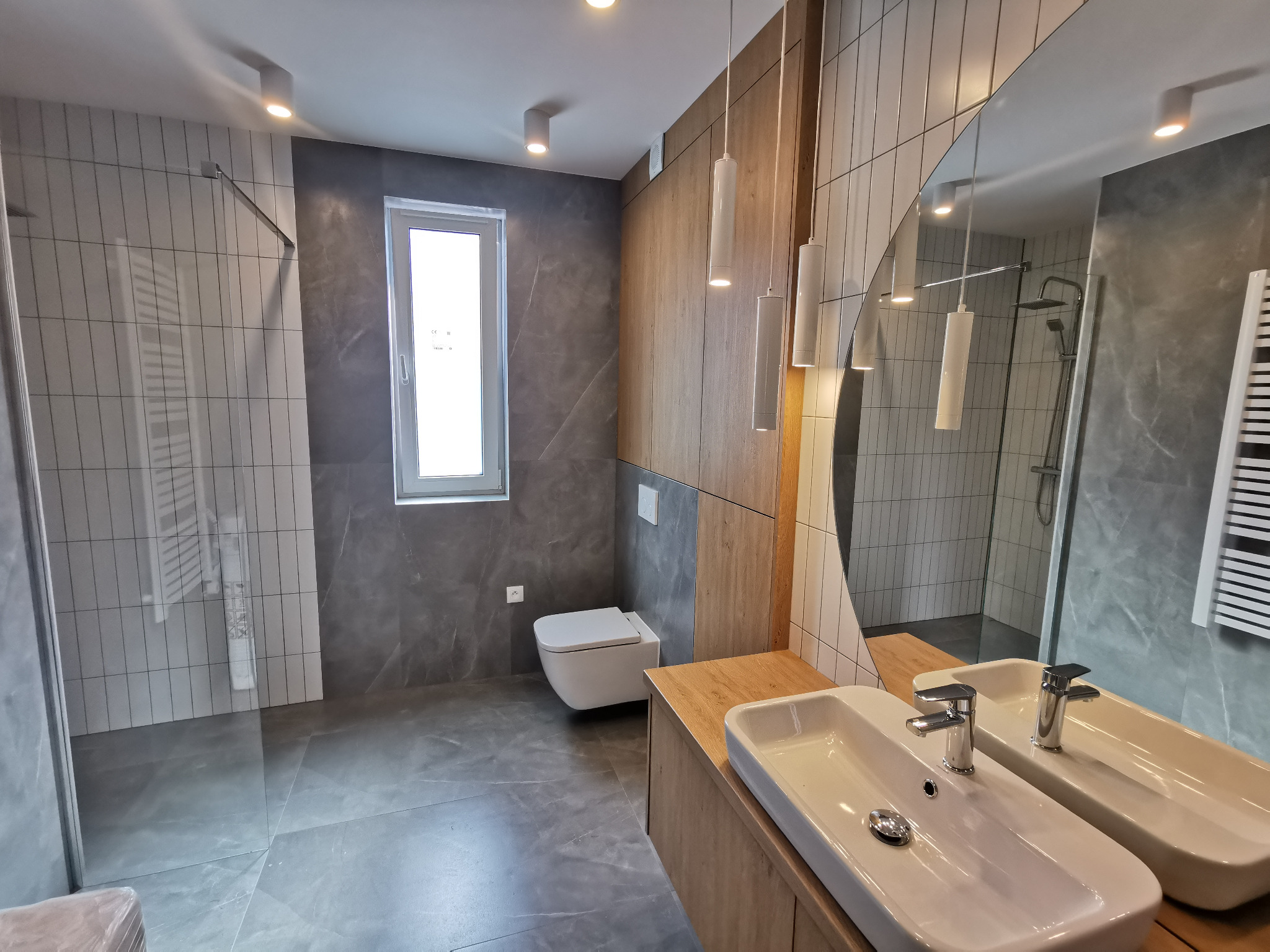 Nowoczesna łazienka po remoncie: szare płytki imitujące marmur, drewniane akcenty, okrągłe lustro i podwieszana toaleta. Widoczny prysznic typu walk-in.