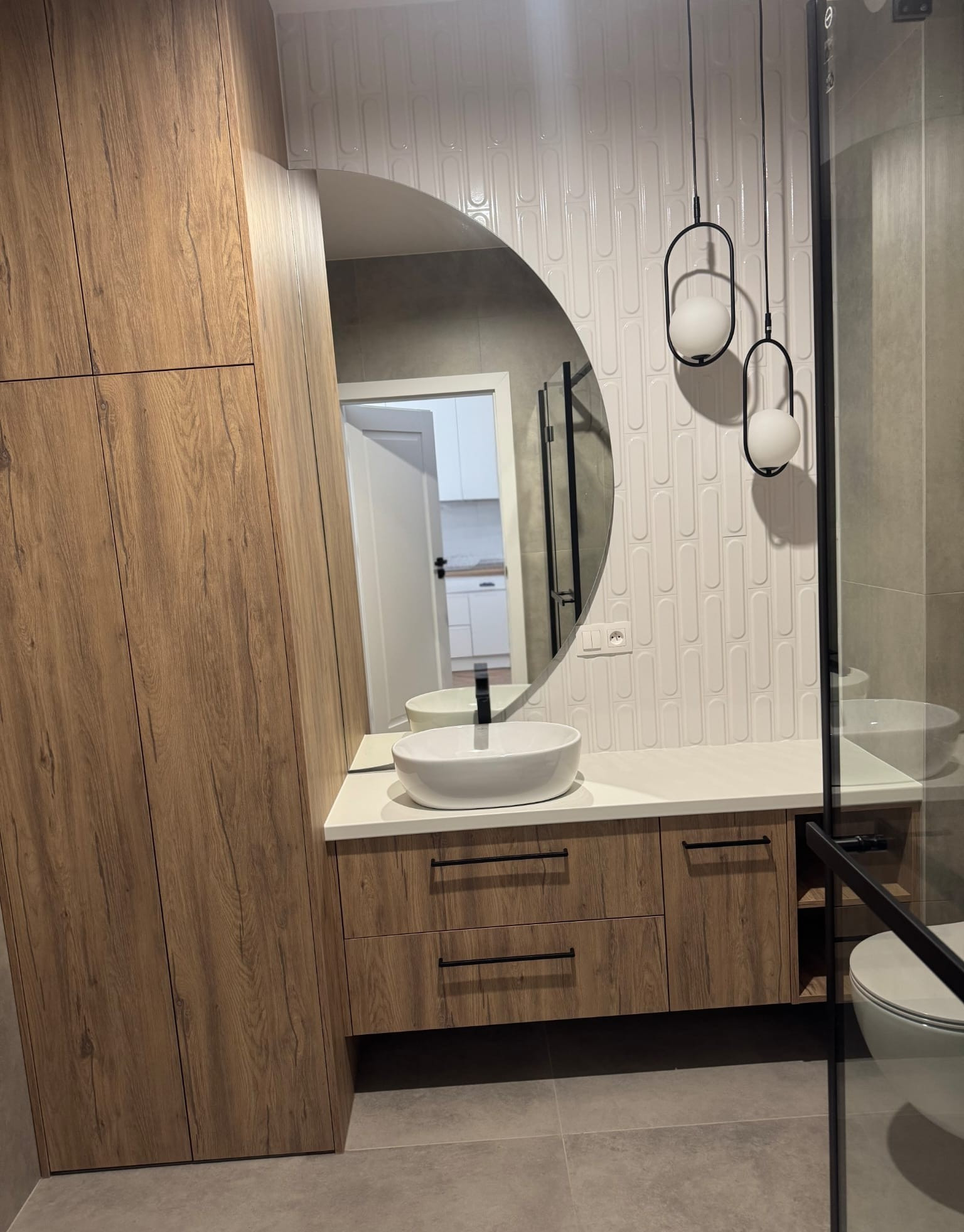 Nowoczesna łazienka po remoncie: umywalka na szafce z drewnopodobnymi frontami, okrągłe lustro i dekoracyjne lampy wiszące. Białe płytki na ścianie.