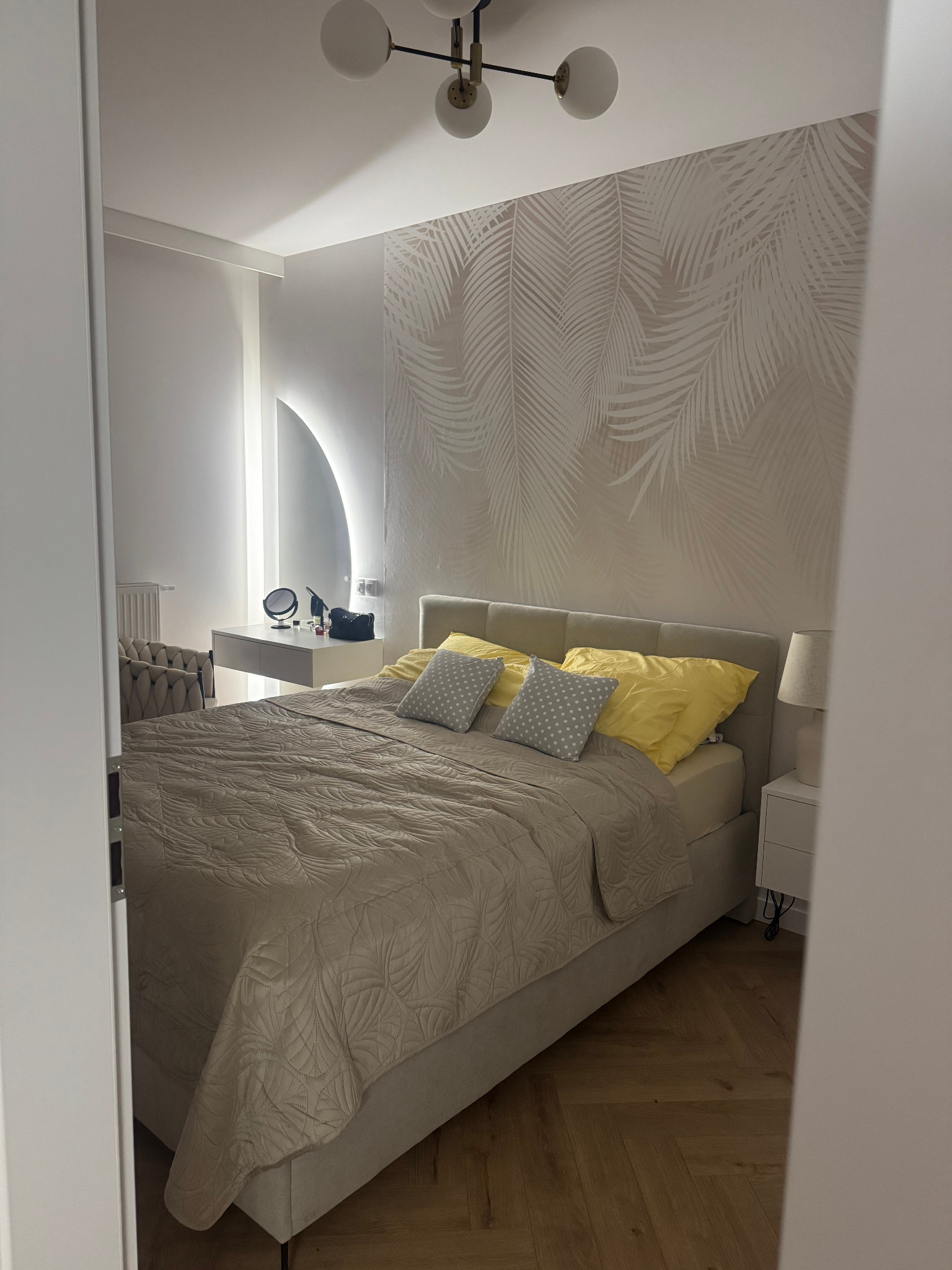 Nowoczesna sypialnia z łóżkiem, tapetą w liście palmowe i toaletką z lustrem LED. Parkiet w jodełkę na podłodze. Widok zza framugi drzwi.