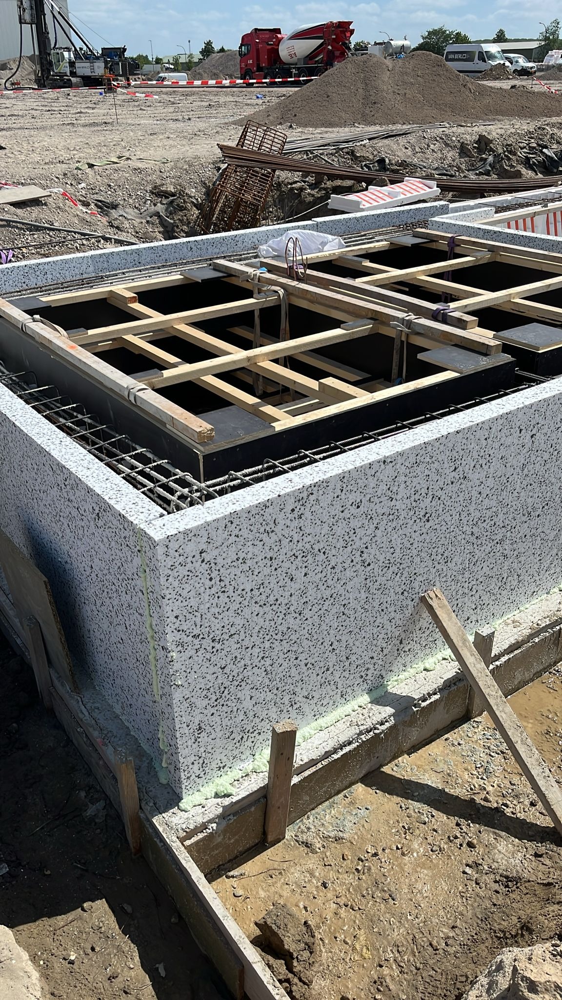 Fundament z bloczków betonowych, wzmocniony zbrojeniem i drewnianym szalunkiem. W tle teren budowy z ciężkim sprzętem i stertą piasku. Widok z góry.