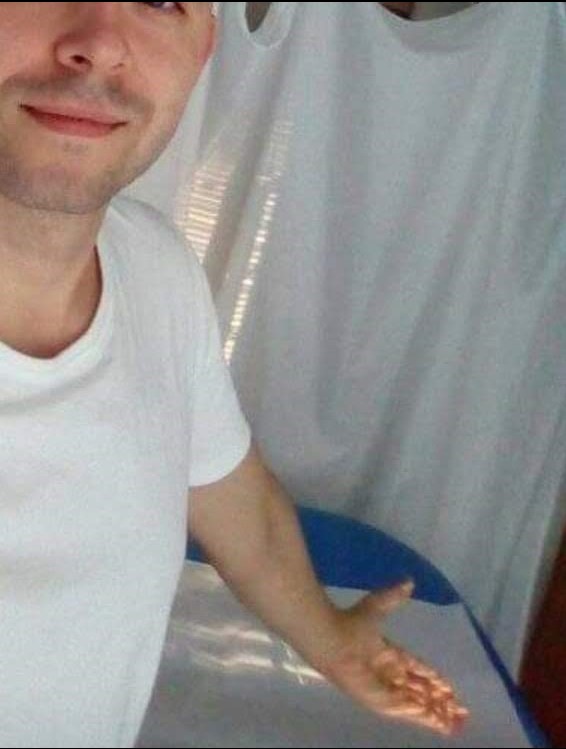 Uśmiechnięty mężczyzna w białej koszulce, z wyciągniętą dłonią na tle białego parawanu i niebieskiego elementu, prawdopodobnie stołu do masażu.