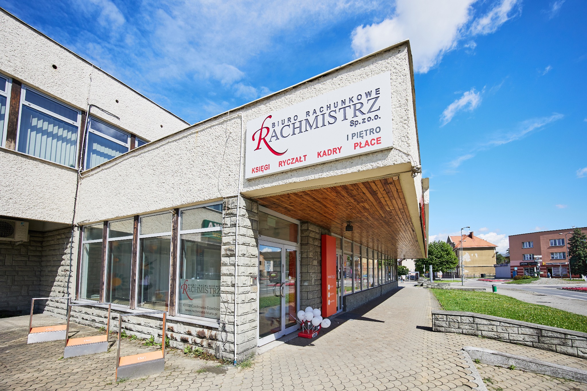 Fasada Biura Rachunkowego Rachmistrz z wyraźnym szyldem informacyjnym, widoczna z poziomu chodnika w słoneczny dzień. Elementy architektury i otoczenia miejskiego.