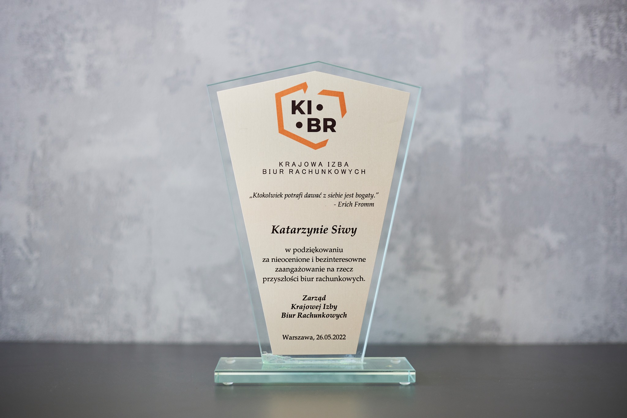 Szklana statuetka z podziękowaniem dla Katarzyny Siwy od Krajowej Izby Biur Rachunkowych, z logo KI•BR, na szarym tle. Wręczona w Warszawie, 26.05.2022.