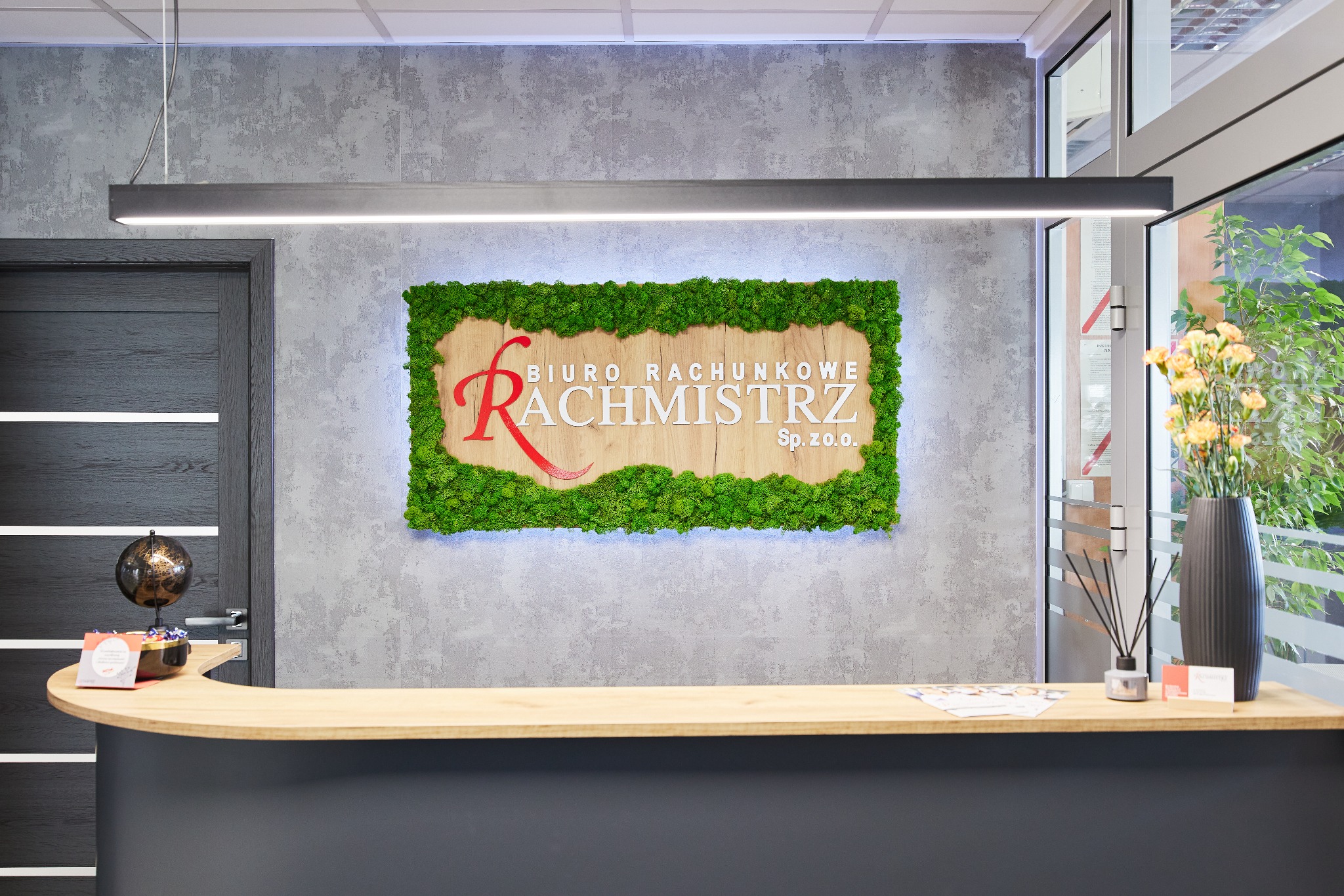 Recepcja biura rachunkowego z logo 'Rachmistrz' otoczonym mchem, podświetlona lampą. Na blacie wazon z kwiatami i ulotki. Nowoczesny design.