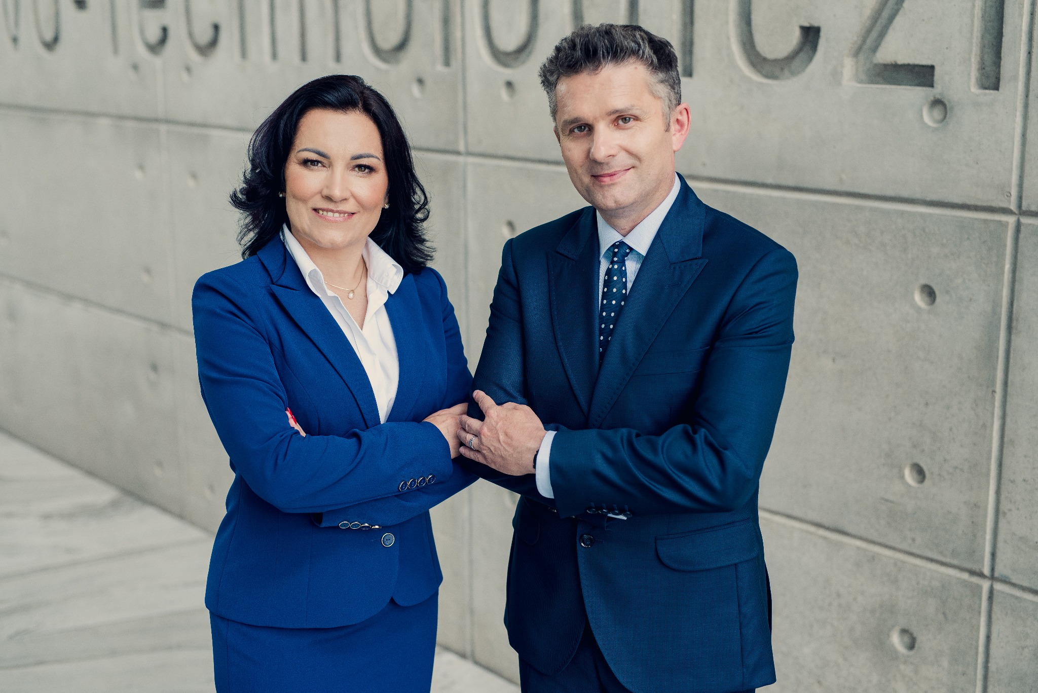 Dwoje uśmiechniętych profesjonalistów w garniturach pozuje na tle betonowej ściany z napisem. Kobieta w niebieskim kostiumie, mężczyzna w granatowym garniturze.