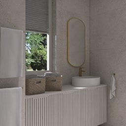 Wiktoria Nowocień NOVO - Jasna łazienka z owalnym lustrem w złotej ramie, okrągłą umywalką i szafką z pionowymi żłobieniami. W oknie rolety i widok na zieleń. Dwa kosze z ręcznikami.